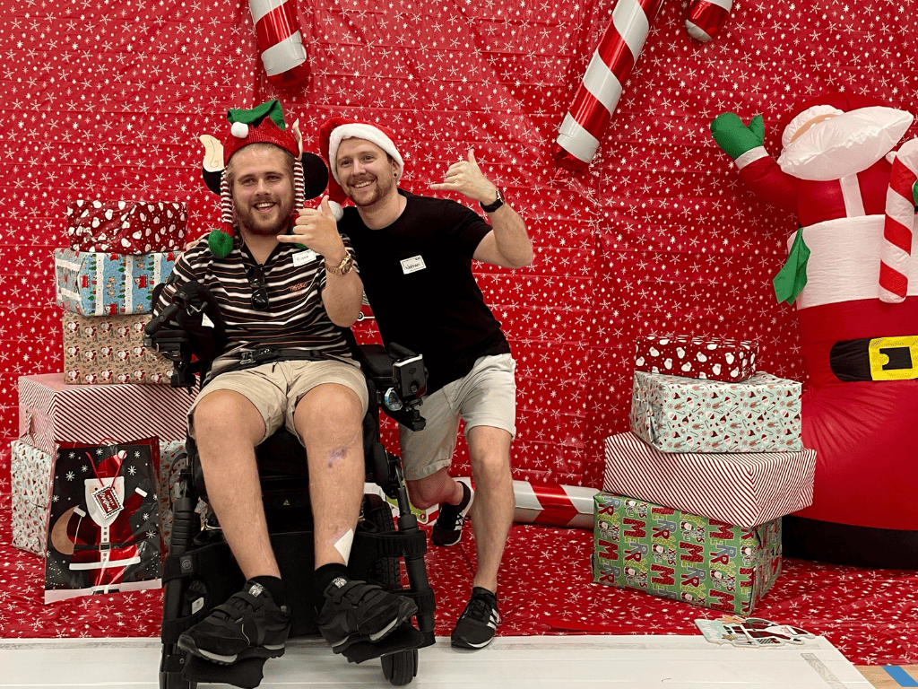 Exercise Physiologist Nathan with Sporting Wheelies member in a wheelchair. Both are smiling at the camera and they are surrounded with Christmas decorations