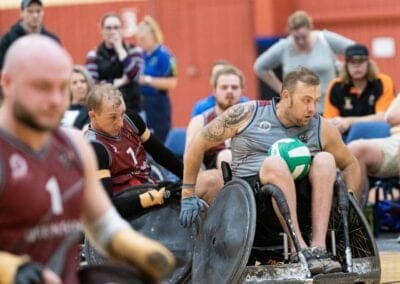 A male wheelchair rugby player rolls down the court with a ball in his lap.
