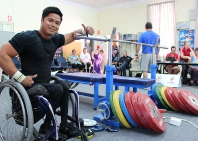 Wheelchair power lifter poses next to bench press equipment at a competition.